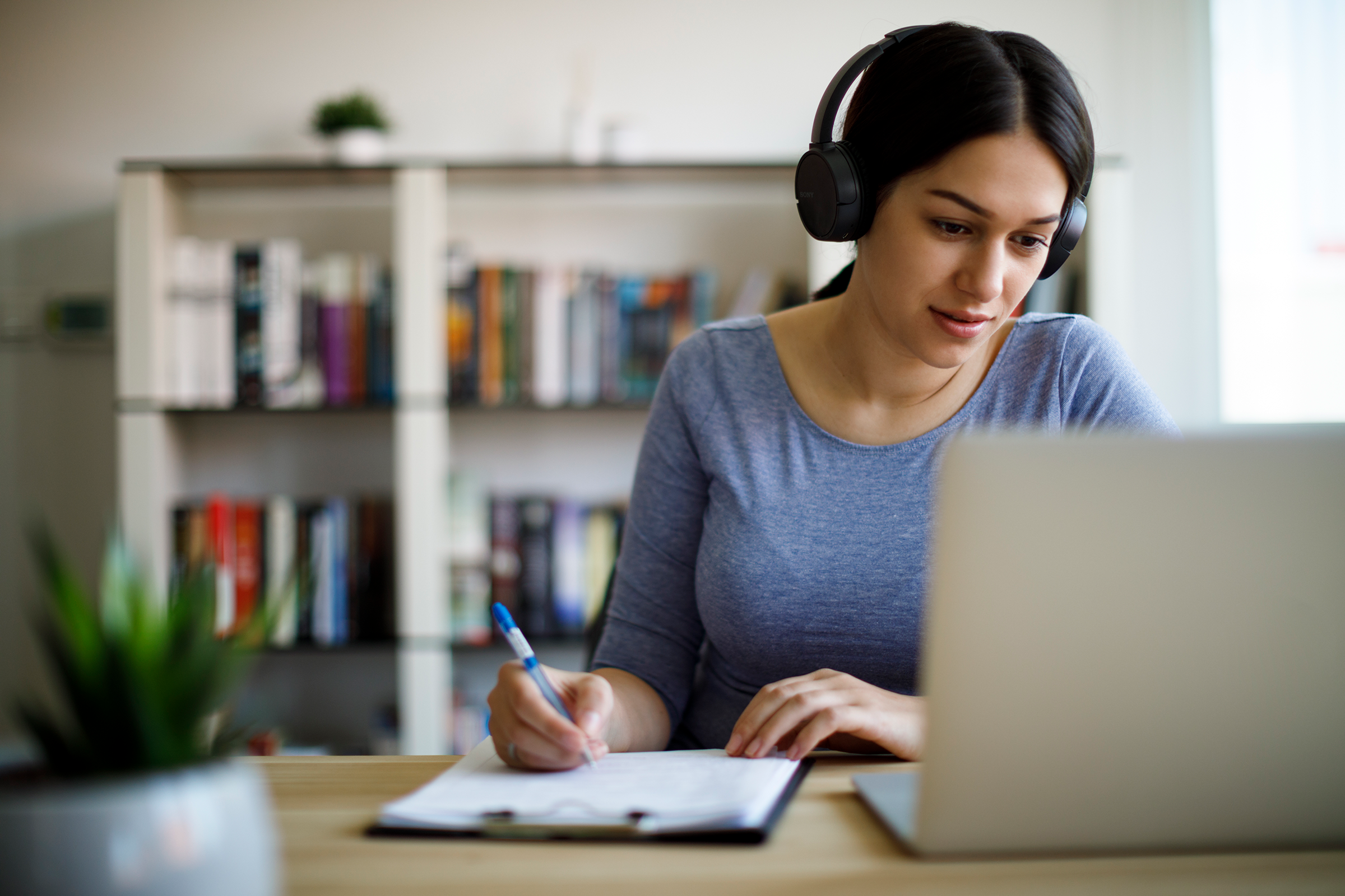 Hispanic woman wearing headphones and looking at computer in a home office setting.