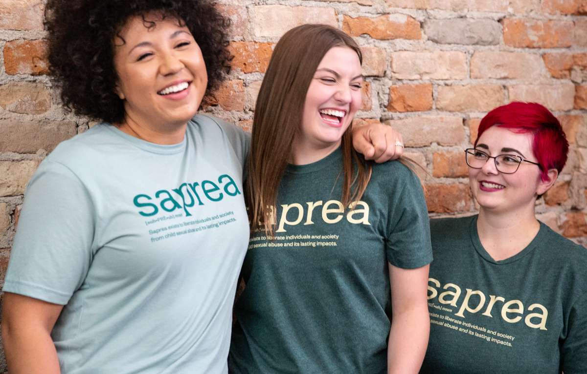 three women standing together against a brick wall wearing Saprea t-shirts. Two are wearing a dark green and one a light green t-shirt with the Saprea logo and mission statement imprinted on them.