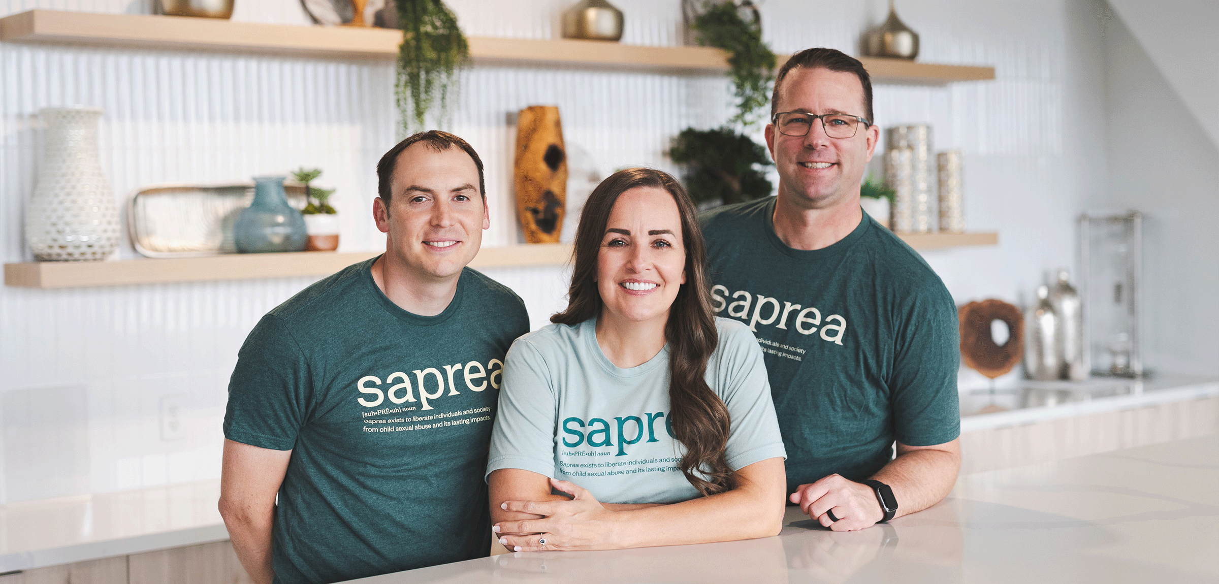 Three people wearing matching "Saprea" t-shirts smile while posing together in a modern, light-filled space with shelves decorated with plants and ceramics.