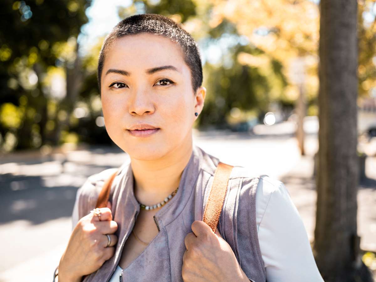 A confident woman with a buzz cut stands outdoors on a sunny day, looking directly at the camera. They are wearing a light-colored shirt and a gray vest, with a beaded necklace and small earrings. A backpack strap is visible over each shoulder, and the background is softly blurred with trees and a street scene.