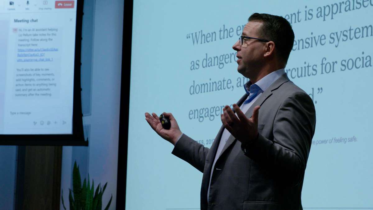 Middle aged man presenting to a group standing in front of a projection screen. He is looking out to crowd with his arms open.